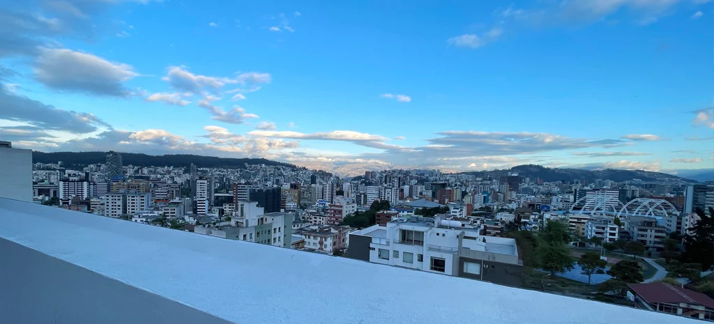 Fotografía de la terraza de Kamay: borde de muro blanco con vista a la ciudad, a cerros y a un cielo con nubes.