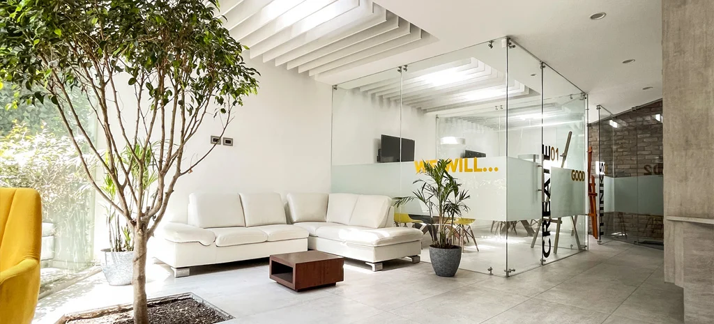 Fotografía de Kapía: sala con sofá blanco, árbol de interior y salas de vidrio al fondo.