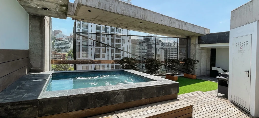 Fotografía de la terraza de Kapía: jacuzzi con borde de piedra, piso de madera y edificios vecinos.