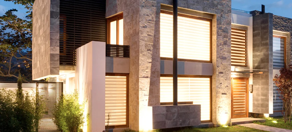 Fotografía de una casa de Domaine III al anochecer: fachadas de piedra, celosías de madera y ventanas iluminadas.