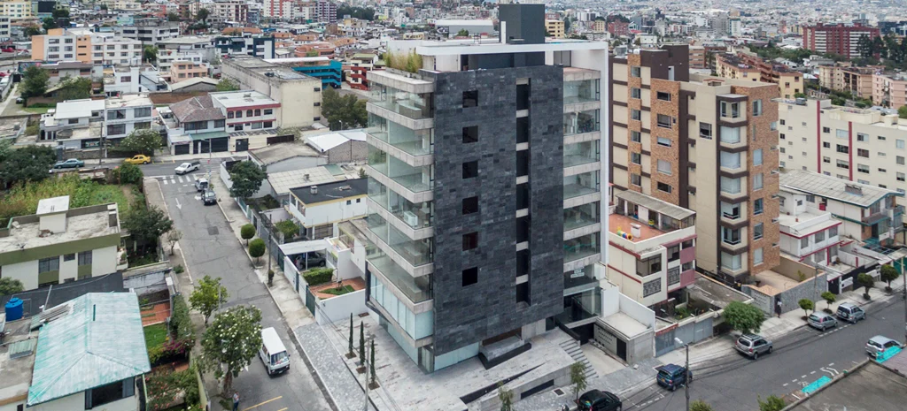 Vista elevada de Kamay: torre de fachada oscura con balcones de vidrio, entre edificios y calles de la ciudad, de día.