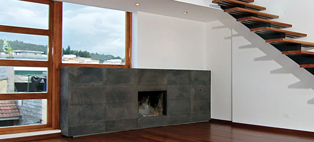 Fotografía de la sala del Edificio Monteserrín: chimenea de revestimiento oscuro, ventanal con marco de madera y escalera.